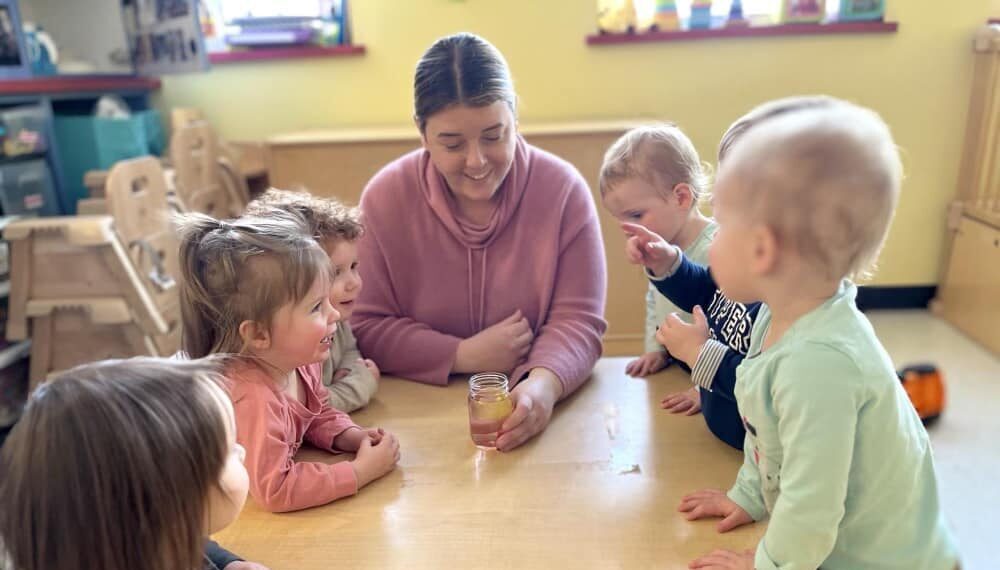 Lakes Region Child Care Services Group of preschool children at a child-size wooden table conversing animatedly and pointing while a smiling teacher shows them a jar with liquid in it