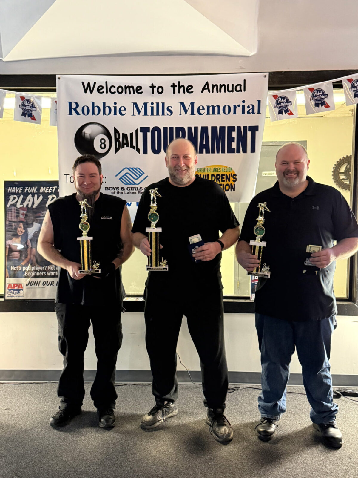 3 winners of the Robbie Mills pool tournament holding trophies in front of the event banner